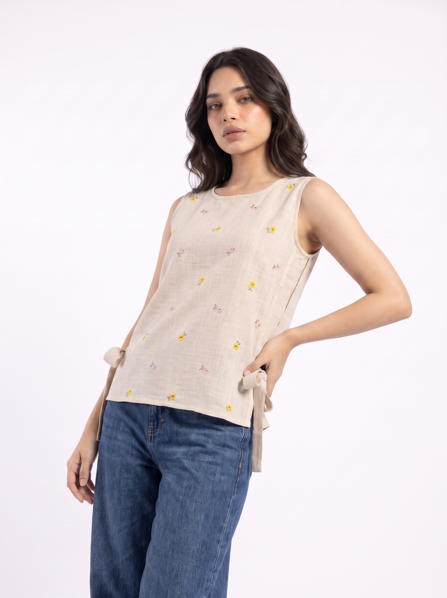 A woman modeling a sleeveless beige linen top with a small yellow and pink floral print, paired with blue jeans, against a plain white studio background.