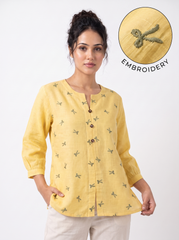 A woman wearing a yellow linen tunic top with small embroidered motifs, paired with beige trousers, against a plain white background. A circular inset in the top right highlights the embroidery detail with the word 'EMBROIDERY'.