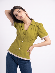 A woman modeling a short-sleeve olive green blouse with cream-colored ruffle trim at the collar, cuffs, and hem, paired with blue jeans against a plain white studio background.