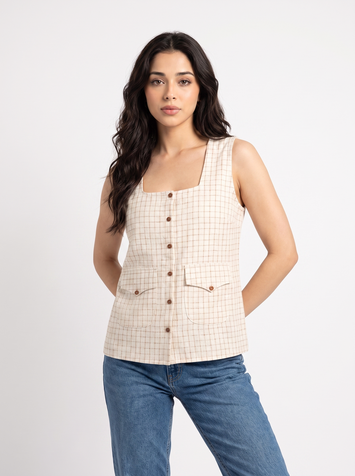 A woman modeling a sleeveless beige and white checkered vest with brown buttons, paired with blue jeans, against a plain light gray background.