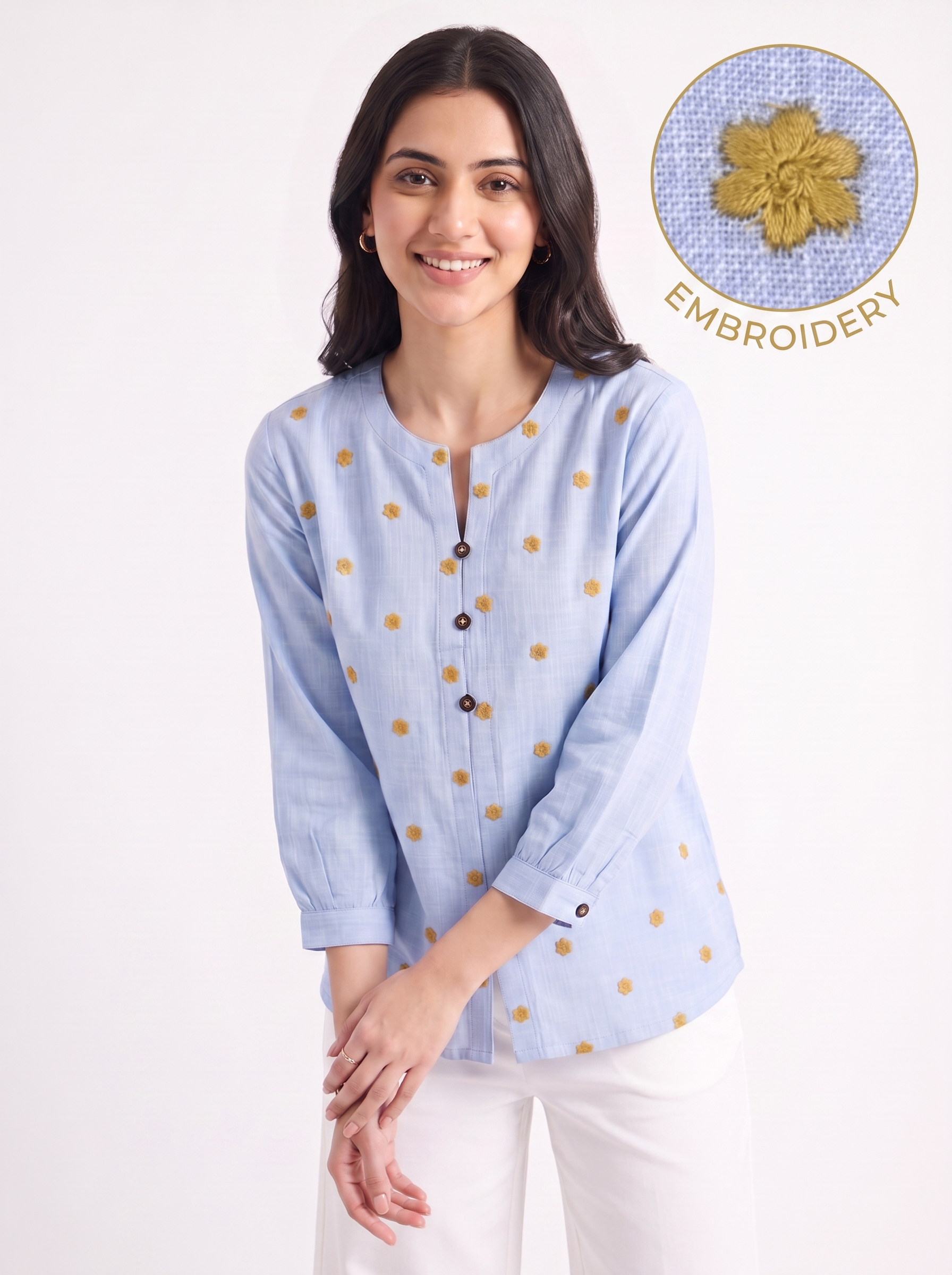 A woman wearing a light blue long-sleeve top with small gold embroidered dots, paired with white pants, against a plain white background. An inset circle in the top right shows a close-up of the gold embroidery texture with the word 'EMBROIDERY'.