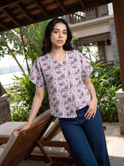 A woman wearing a short-sleeve V-neck blouse with a purple and pink floral pattern on a light background, paired with dark blue jeans, standing in a tropical resort setting.