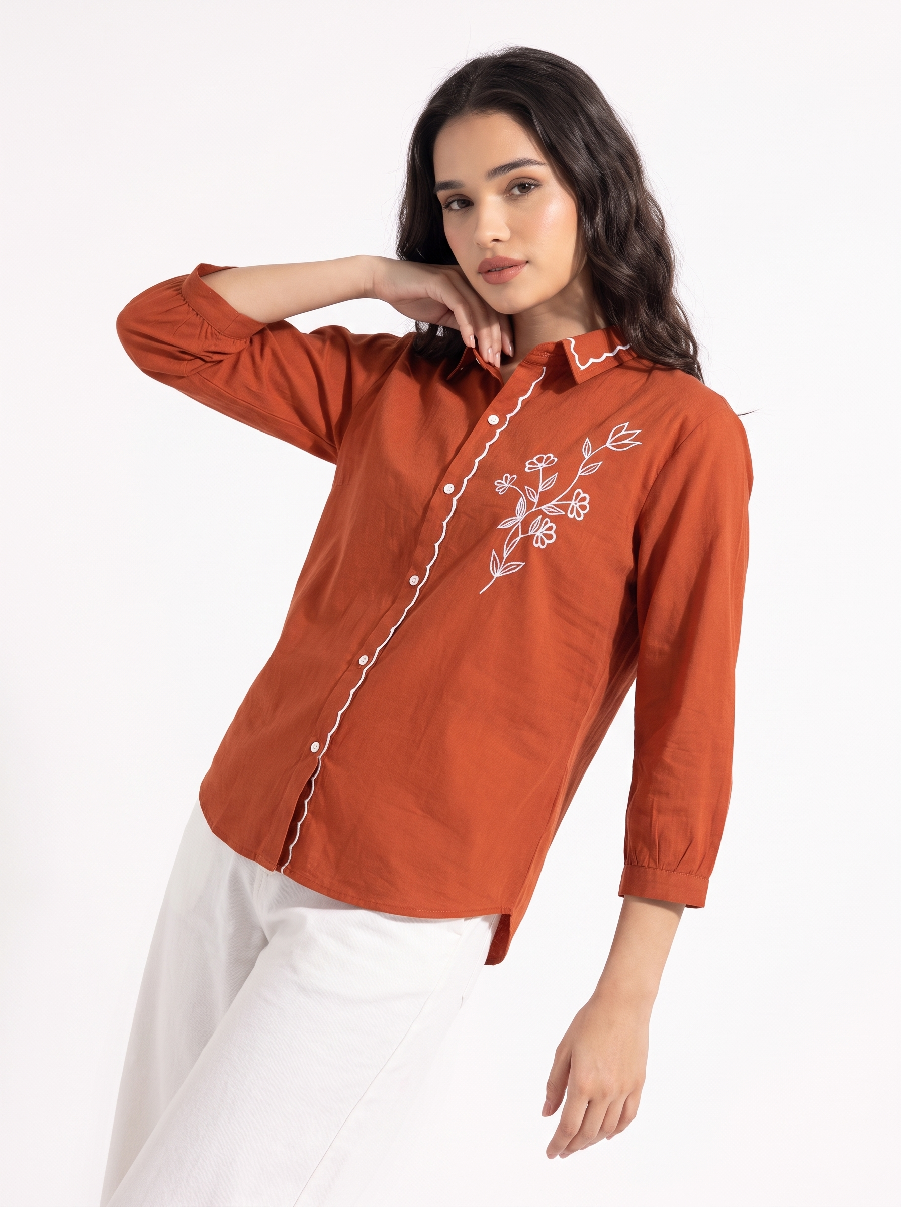 A woman modeling a rust-orange long-sleeve button-up shirt with white floral embroidery on the chest and white piping along the collar and placket, paired with white pants against a plain white studio background.