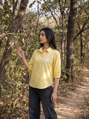 A woman wearing a light yellow embroidered button-up shirt and dark jeans stands on a dirt path in a wooded outdoor setting.