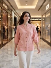 A woman walking down a luxury shopping mall corridor wearing a light pink button-up blouse with small embroidered details and white pants. Gucci and Louis Vuitton storefronts are visible in the background.