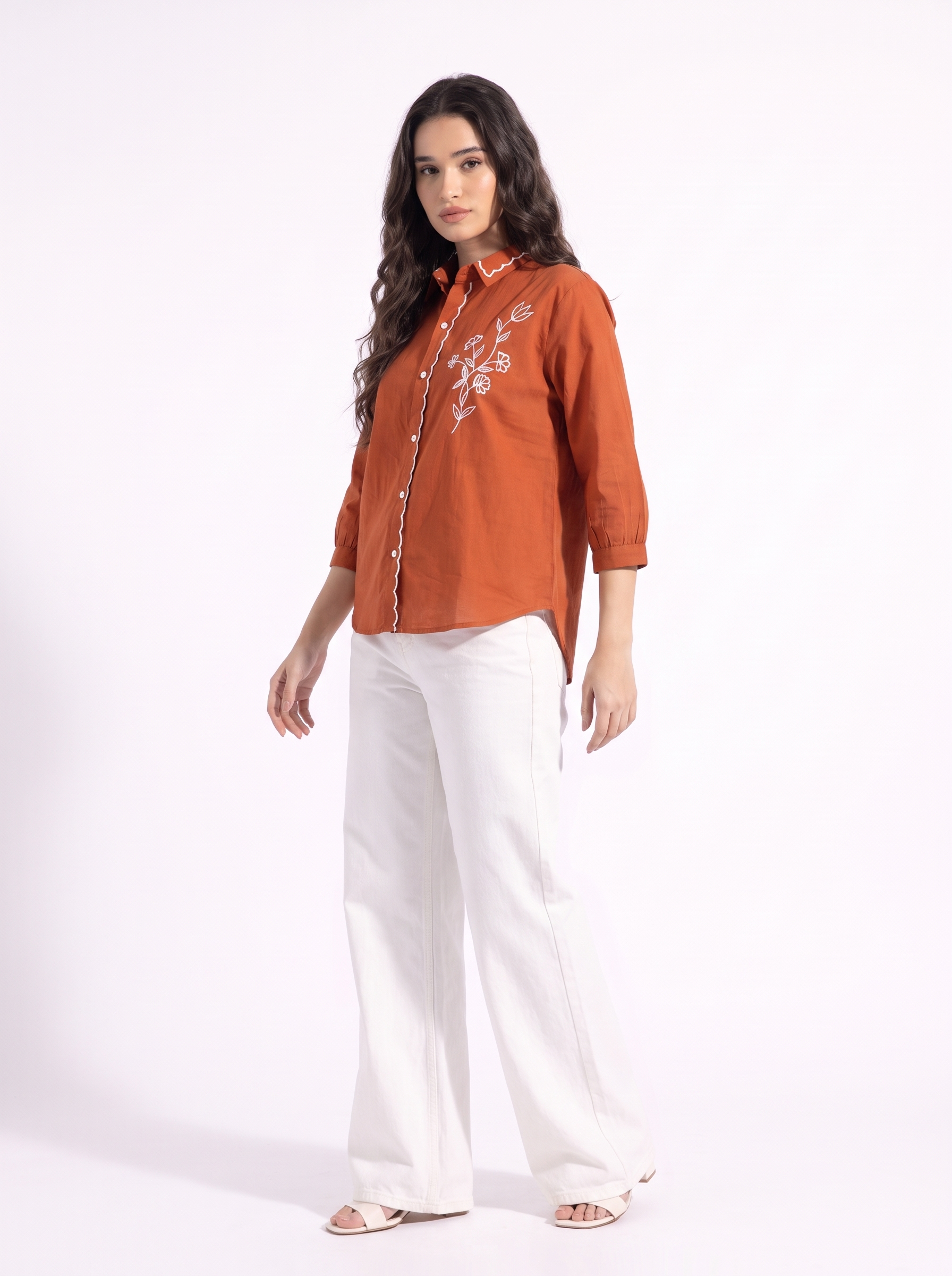 A woman modeling a rust-orange button-up shirt with white floral embroidery on the chest, paired with white wide-leg trousers and beige sandals, against a plain white studio background.