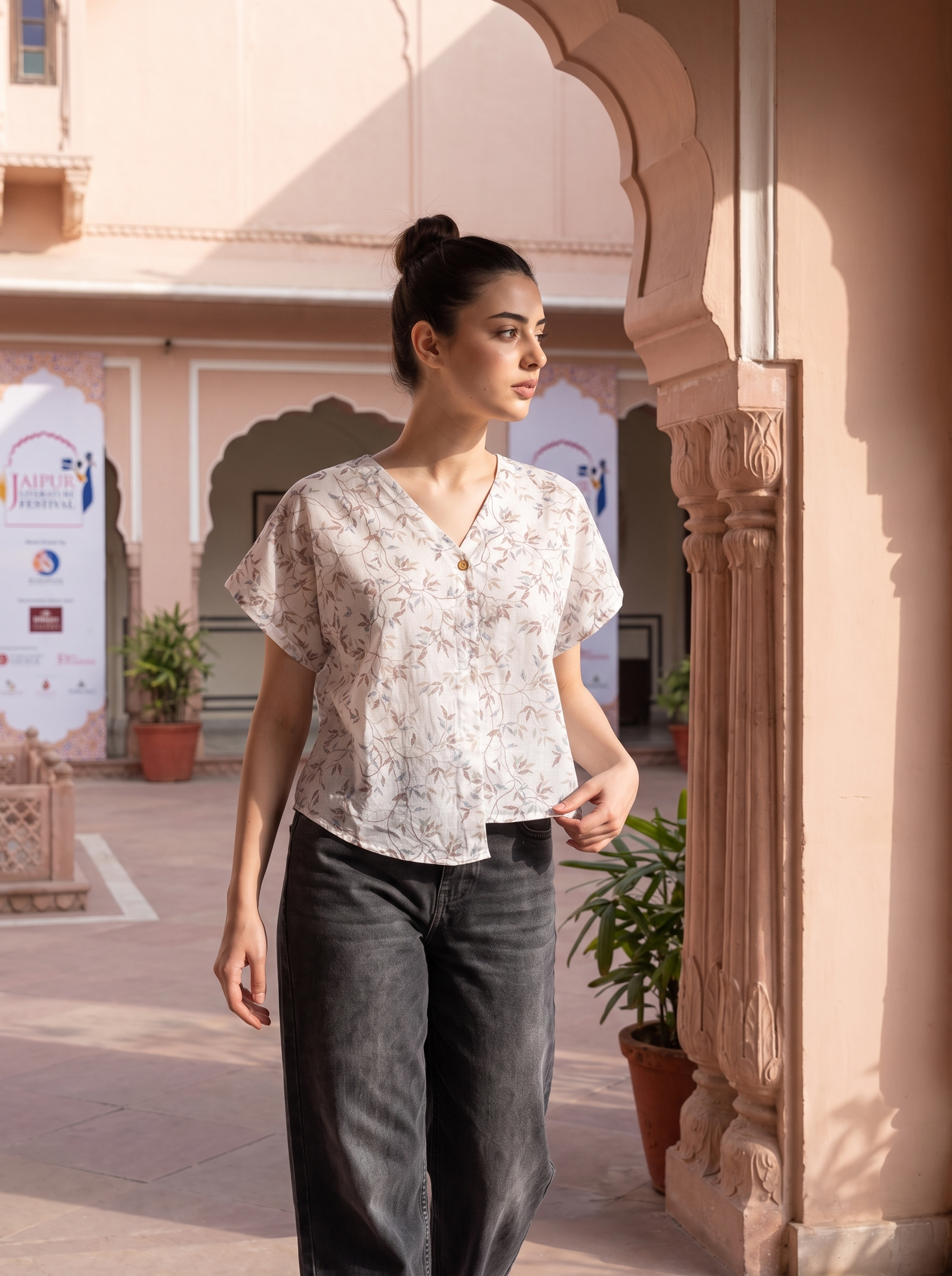 A woman wearing a short-sleeve V-neck blouse with a delicate beige and white floral pattern, paired with dark gray wide-leg jeans, standing in an outdoor courtyard with pink arches.