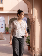 A woman wearing a short-sleeve V-neck blouse with a delicate beige and white floral pattern, paired with dark gray wide-leg jeans, standing in an outdoor courtyard with pink arches.
