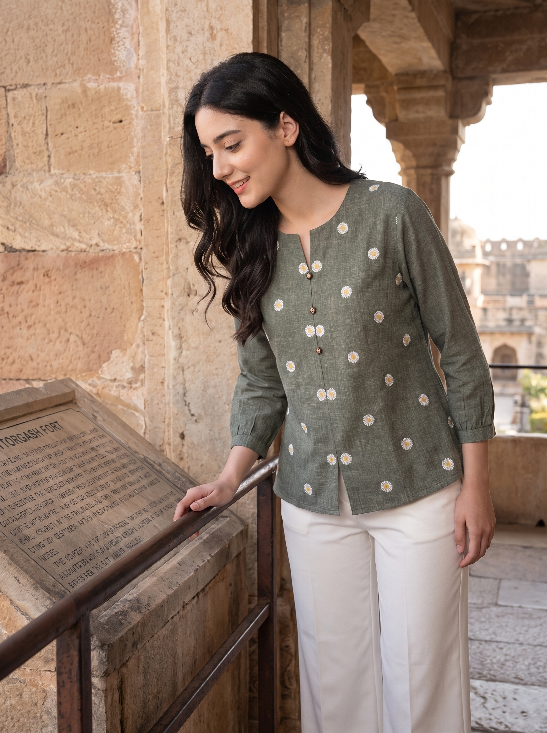 A woman wearing a sage green linen kurta with white polka dots and white trousers, standing in a stone corridor next to a wooden railing.