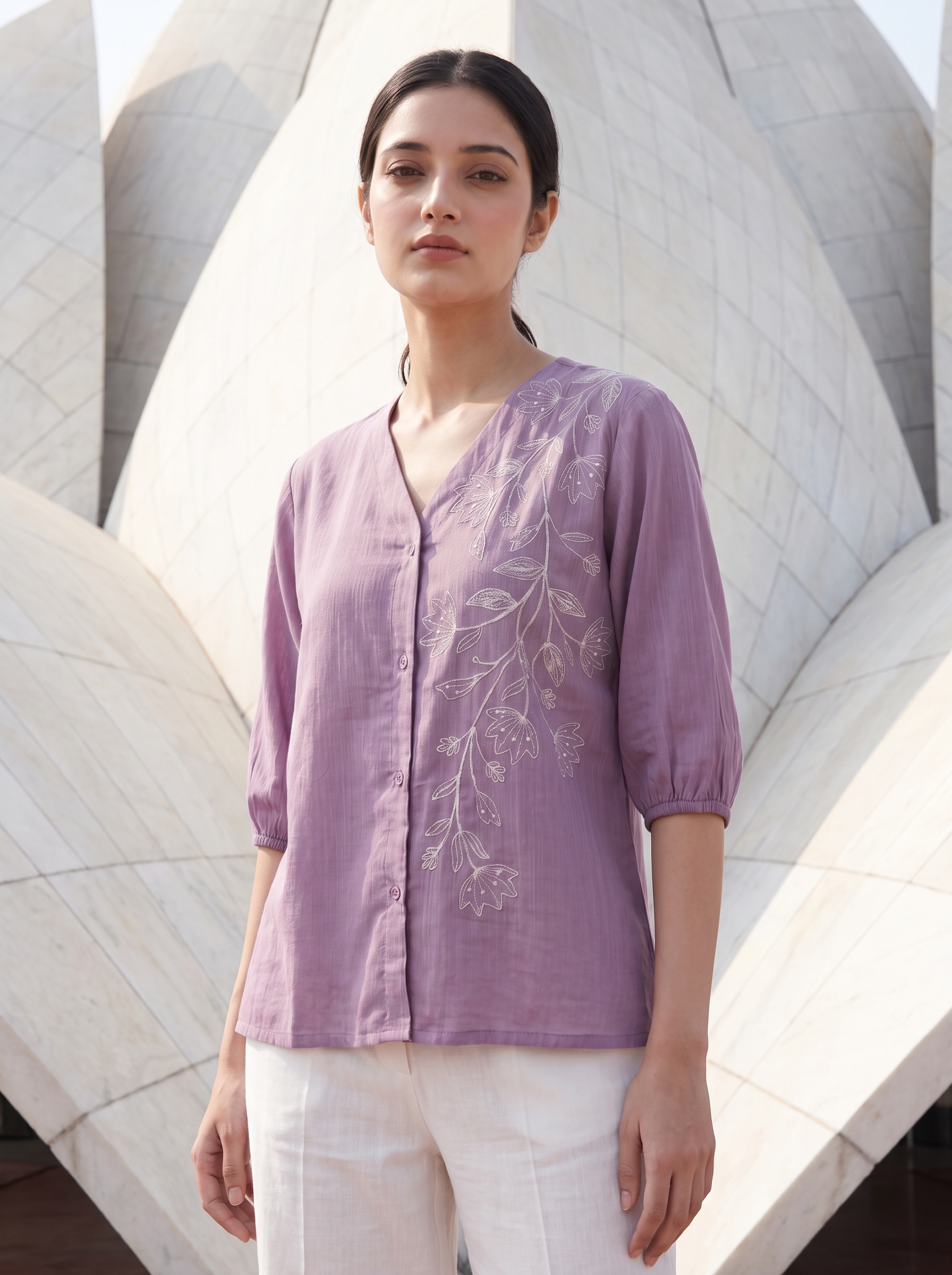 A woman wearing a lavender purple linen button-up blouse with white floral embroidery on the front, paired with white pants, standing in front of a large white architectural structure.