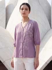 A woman wearing a lavender purple linen button-up blouse with white floral embroidery on the front, paired with white pants, standing in front of a large white architectural structure.