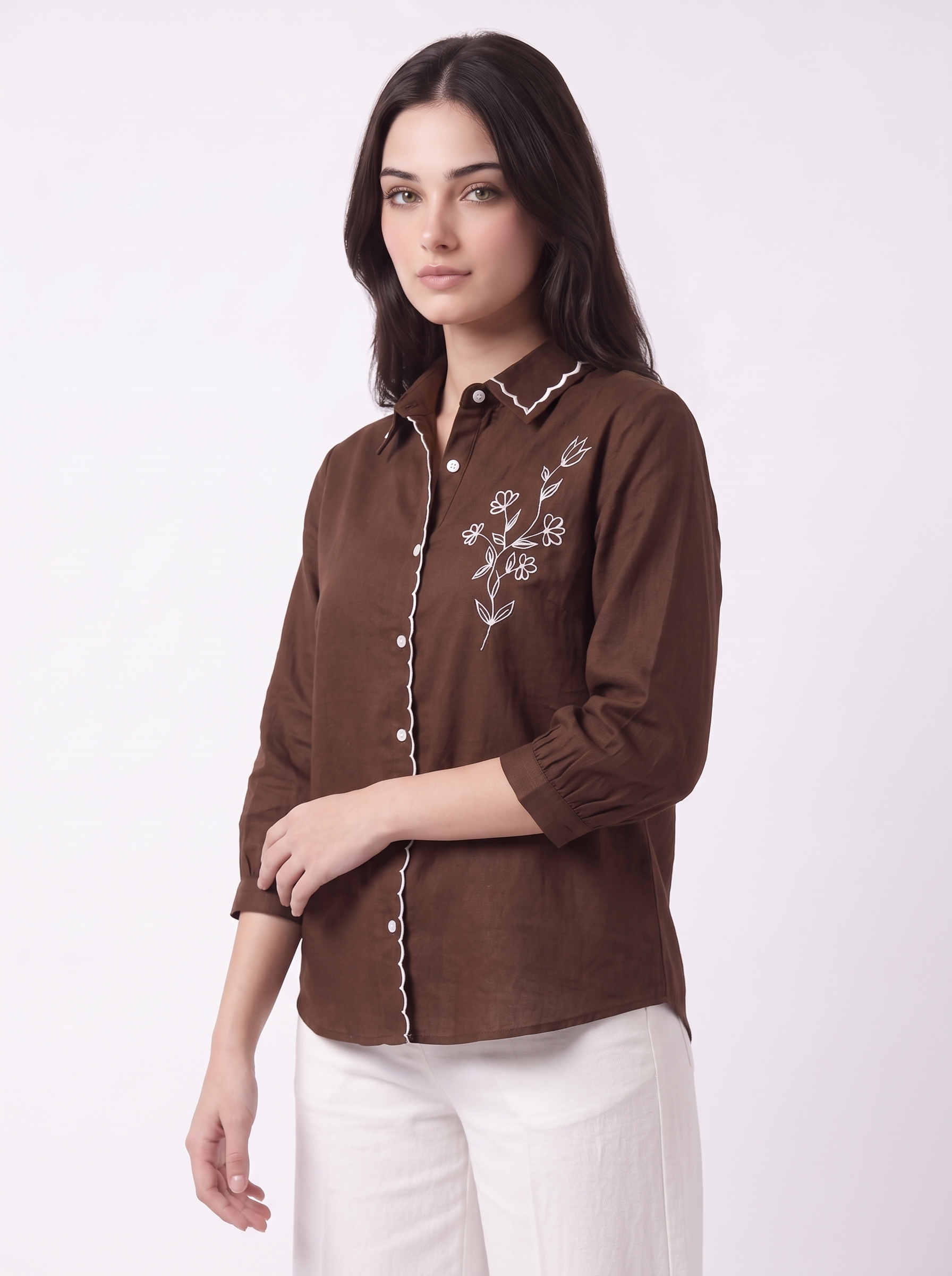 A woman wearing a brown long-sleeve button-up shirt with white floral embroidery on the chest and white pants, photographed against a plain light background.