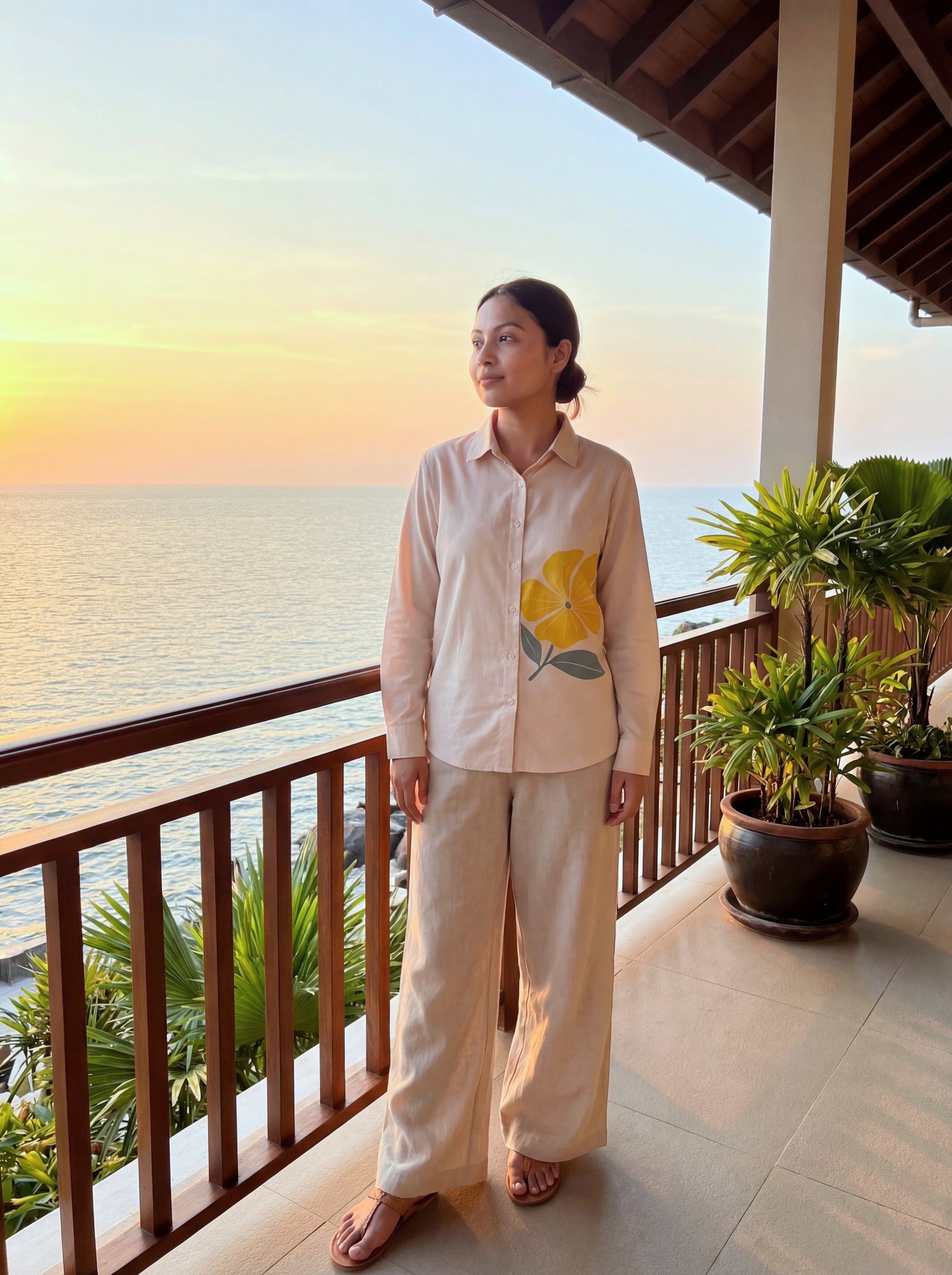 A woman stands on a balcony overlooking the ocean at sunset, wearing a light beige long-sleeve button-up shirt with a yellow floral print and matching wide-leg pants.