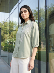 A woman wearing a sage green button-up shirt with white floral embroidery on the chest and white trousers, standing in front of a modern glass building.