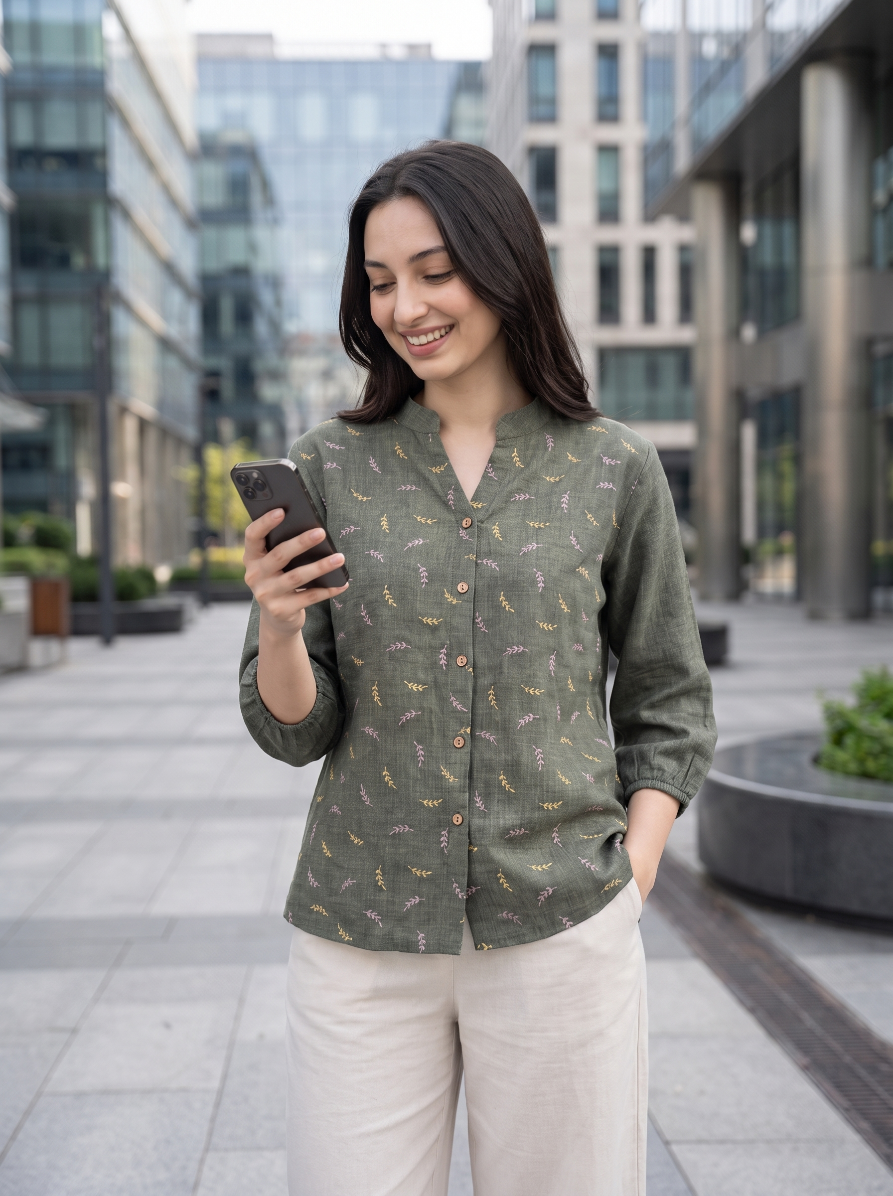 A woman standing outdoors in a modern city plaza, looking at her phone. She is wearing a sage green long-sleeve button-up shirt with a small yellow and white leaf pattern, paired with light beige trousers.