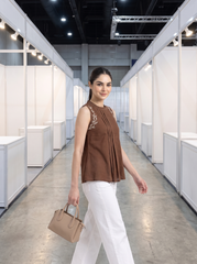 A woman walking down a convention hall aisle wearing a sleeveless brown pleated top with embroidery on the shoulders, white pants, and carrying a beige handbag.