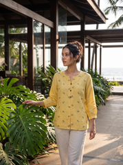 A woman wearing a yellow button-up kurta with small green floral prints and white trousers, standing on a tropical resort walkway next to lush green plants.