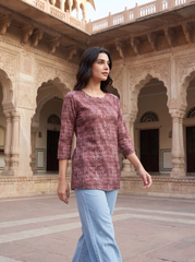 A woman wearing a dusty rose printed kurta with three-quarter sleeves and light blue pants, walking in a courtyard with traditional stone arches.