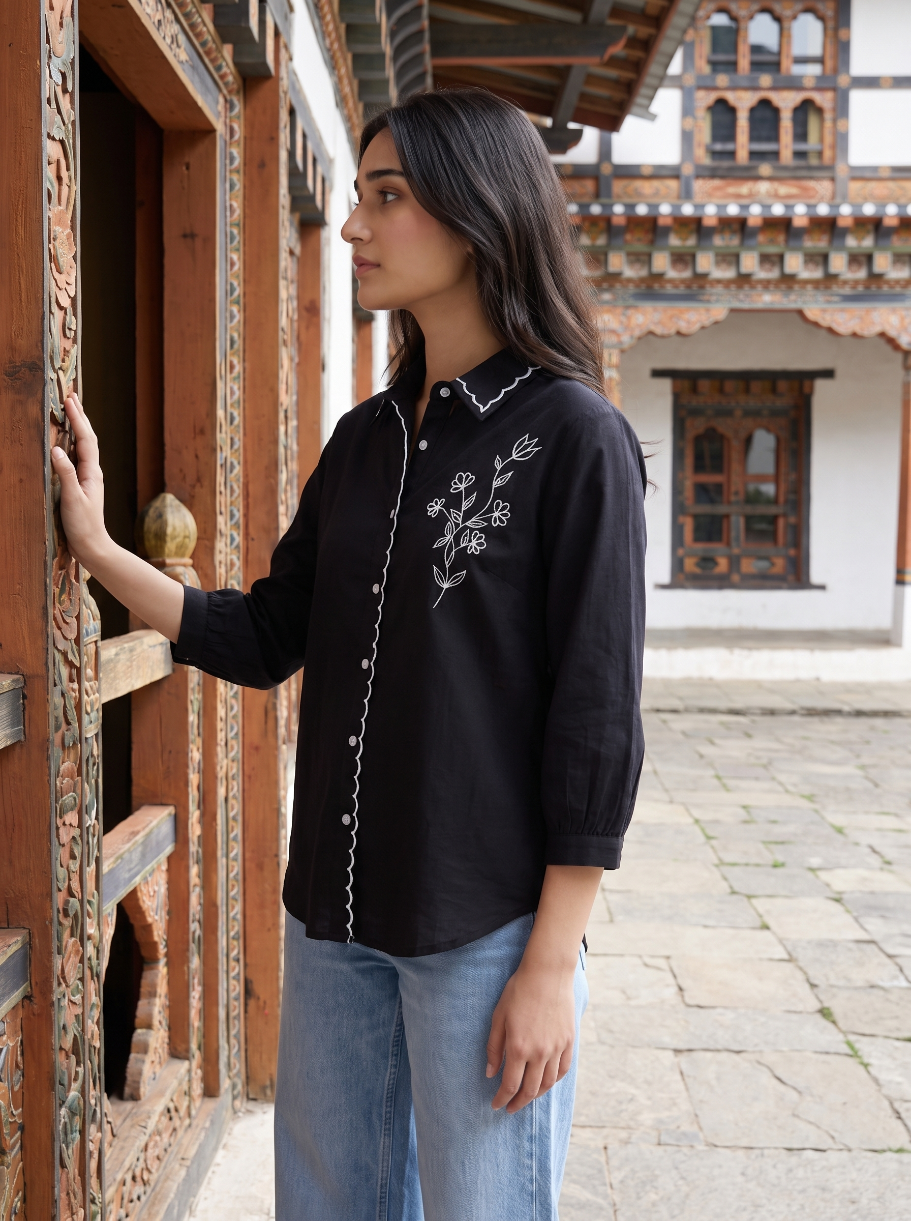 A woman wearing a black long-sleeve button-up shirt with white floral embroidery and white piping, paired with light blue jeans, standing in front of a traditional wooden building.