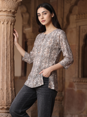 A woman wearing a beige and brown patterned three-quarter sleeve kurta with dark gray jeans, posing next to a carved stone pillar in an architectural setting.