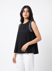 A woman modeling a sleeveless black top with silver-gray embroidered leaf details on the shoulders, paired with white pants, against a plain white studio background.