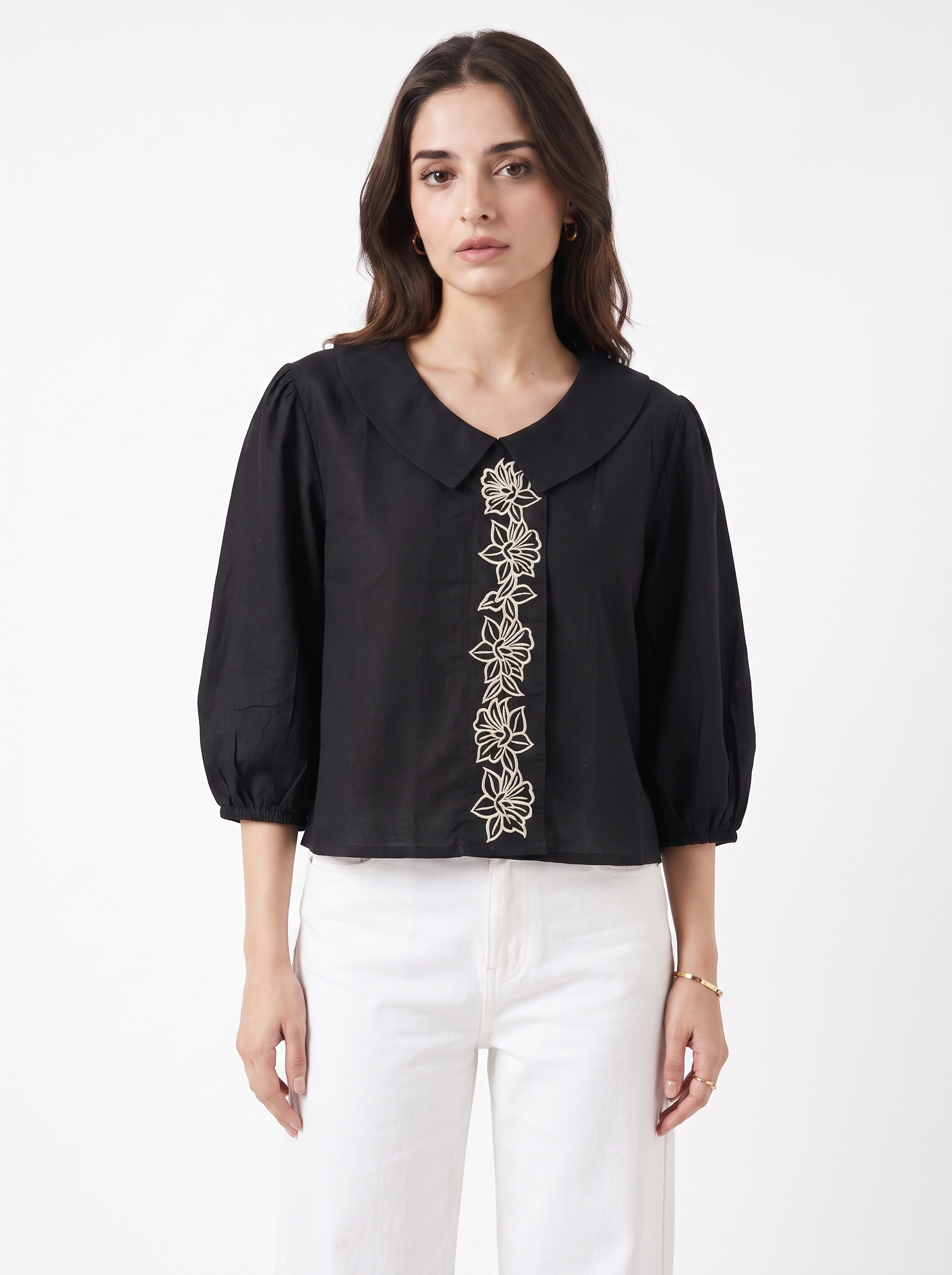 A woman modeling a black long-sleeve blouse with a Peter Pan collar and vertical floral embroidery down the front, paired with white pants, against a plain white studio background.
