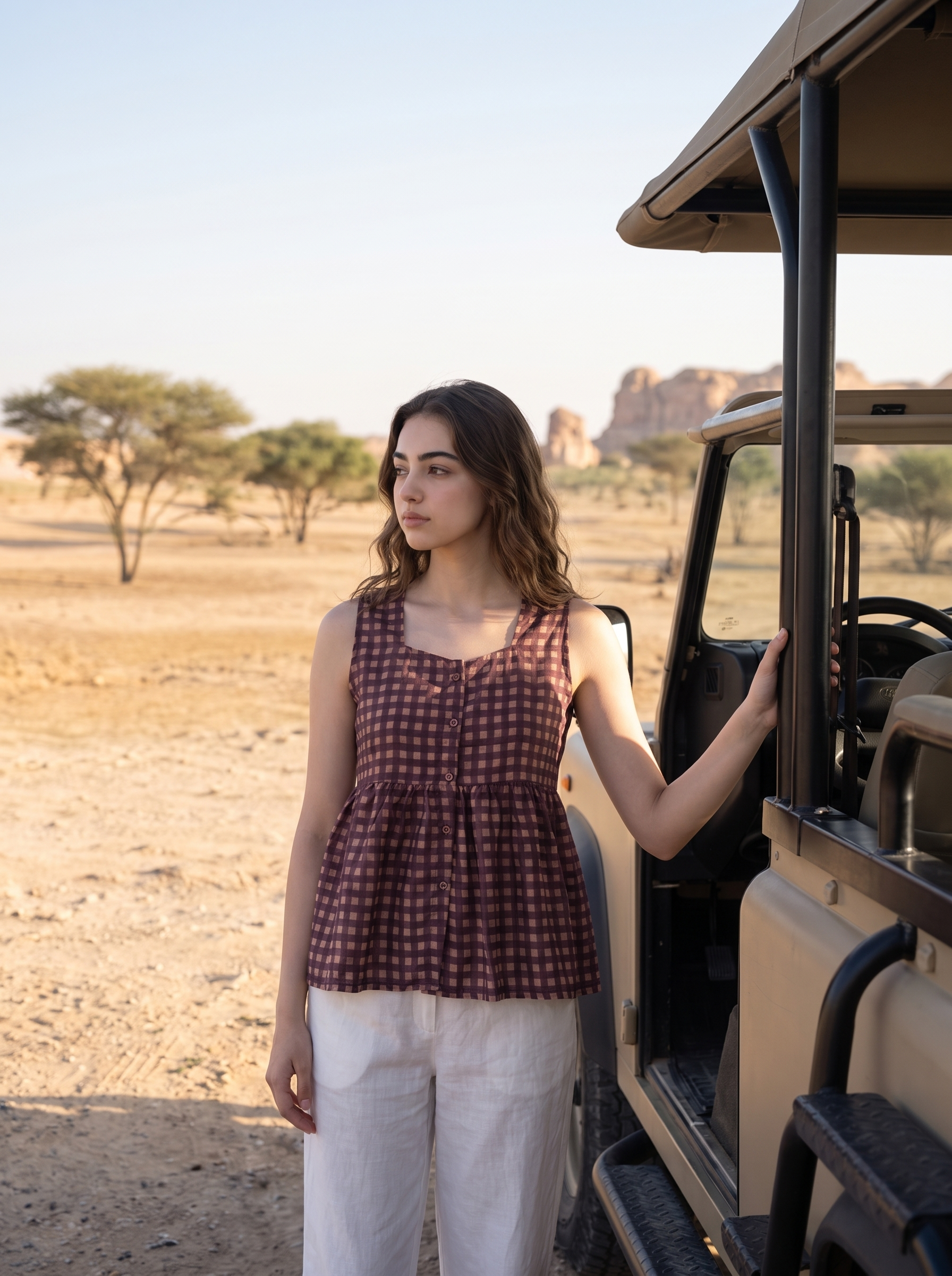 A woman stands beside a safari-style vehicle in a dry desert landscape, wearing a sleeveless burgundy and gold checkered peplum top and white pants.