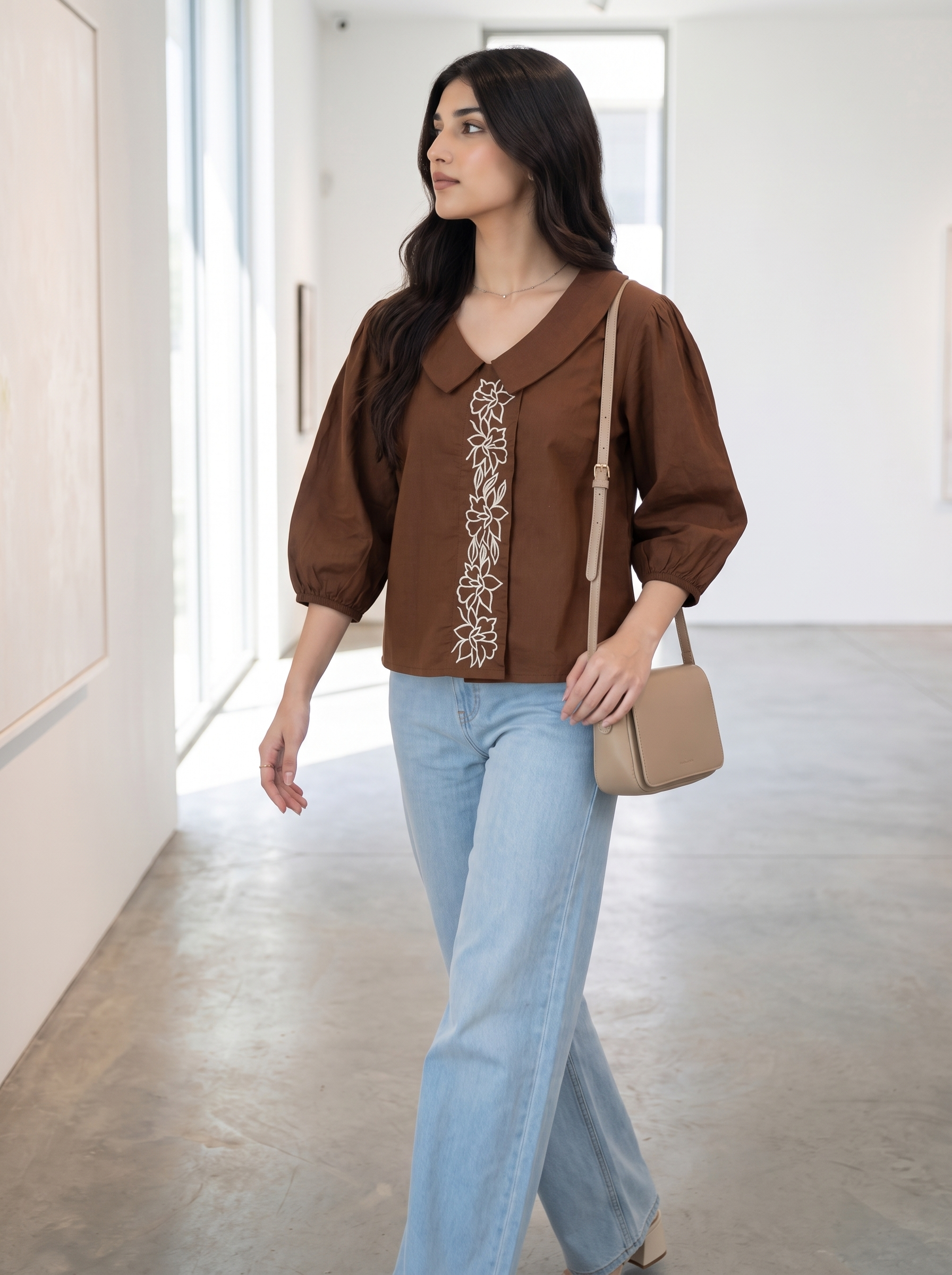 A woman wearing a brown long-sleeve blouse with a decorative white embroidered vertical panel down the front, paired with light blue jeans and carrying a beige crossbody bag, walking in a bright, modern indoor hallway.