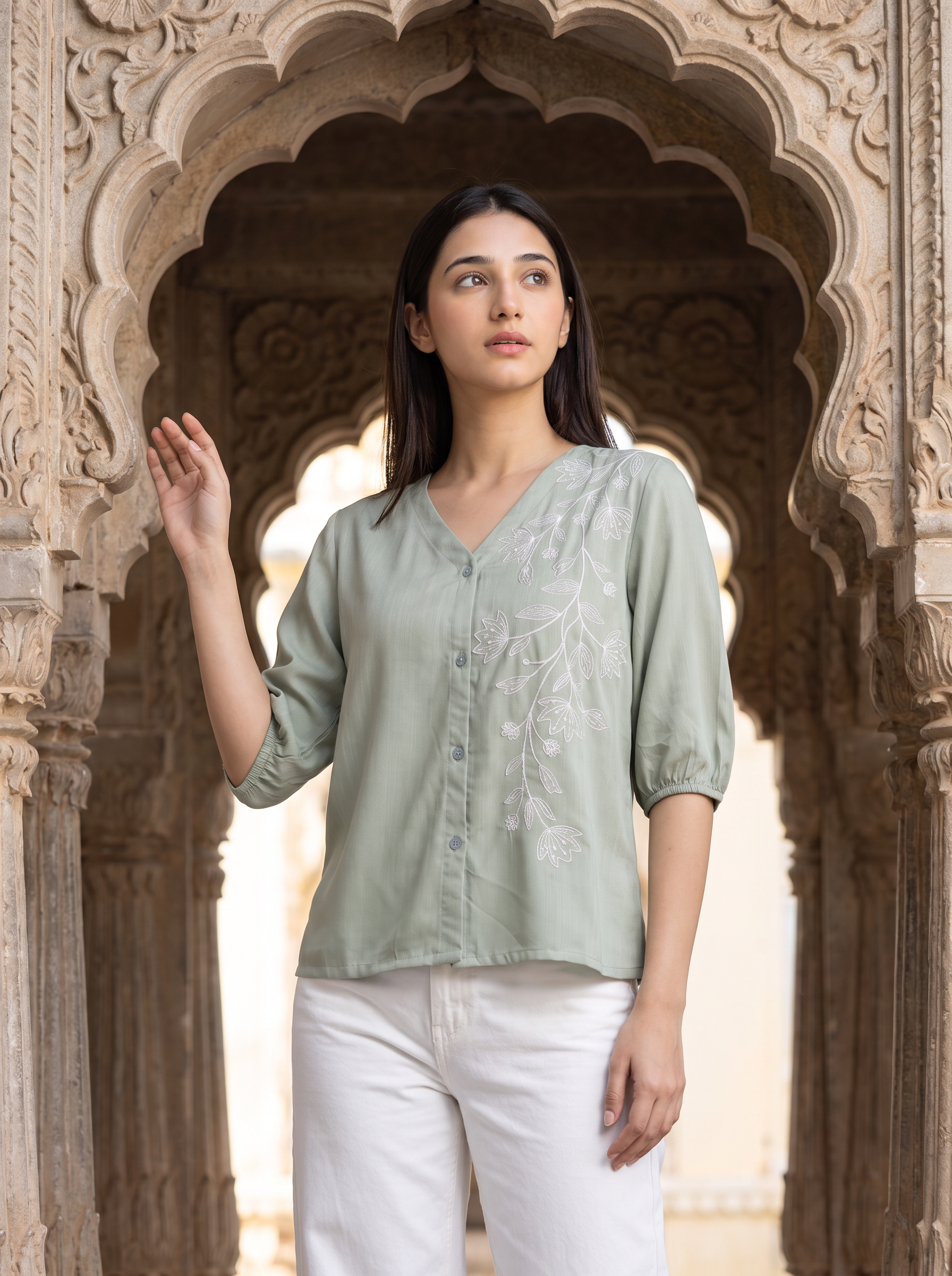 A woman wearing a sage green embroidered blouse and white trousers stands in front of an ornate stone archway.