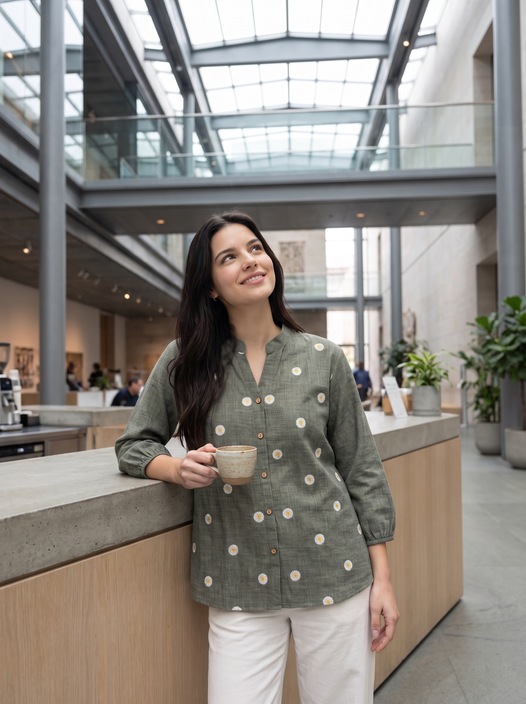 A woman with long dark hair stands in a modern indoor lobby, leaning on a counter and holding a beige ceramic mug. She is wearing a sage green linen button-up shirt with white and yellow polka dots, paired with white pants.