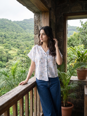 A woman wearing a white short-sleeve blouse with a delicate blue and green floral print, paired with dark blue jeans, walking along a mountain path with misty peaks in the background.