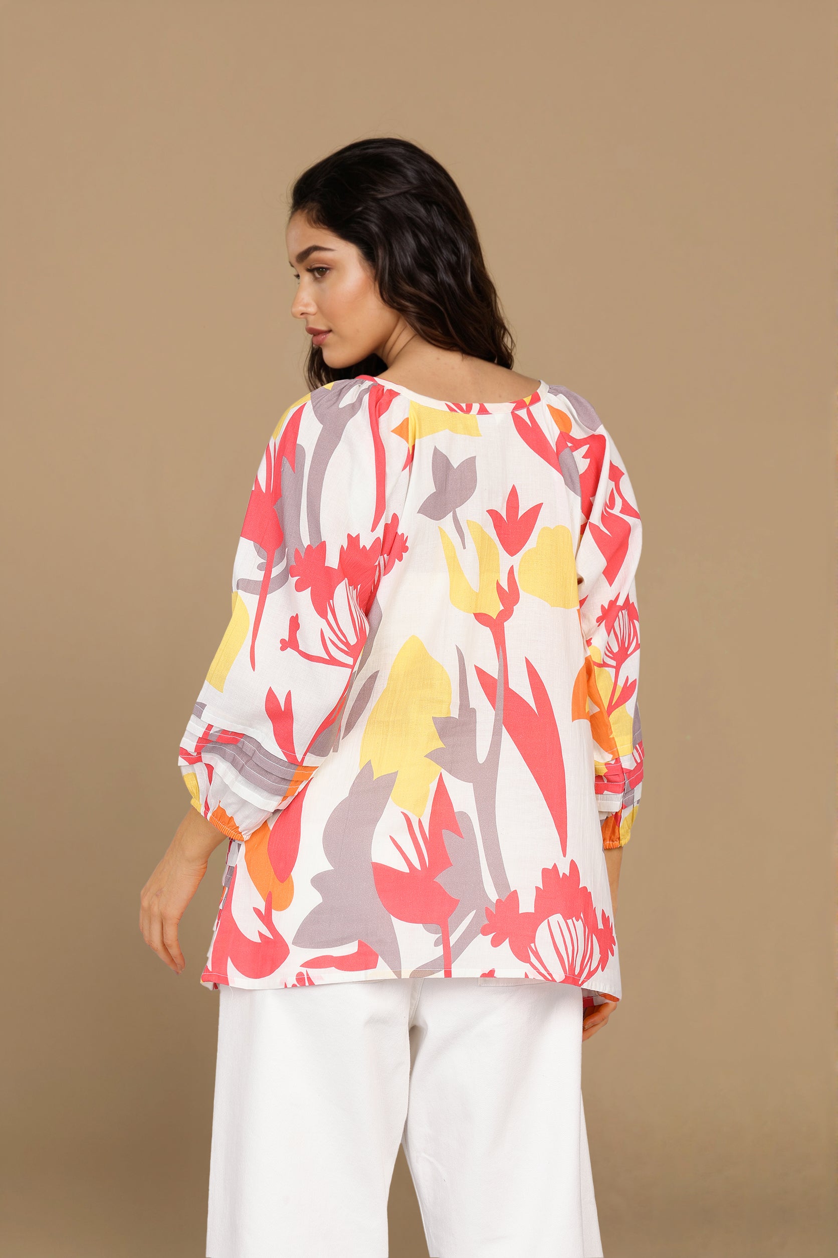 A woman wearing a loose white blouse with a colorful abstract floral print in red, yellow, and gray, paired with white pants, photographed against a solid beige studio background.