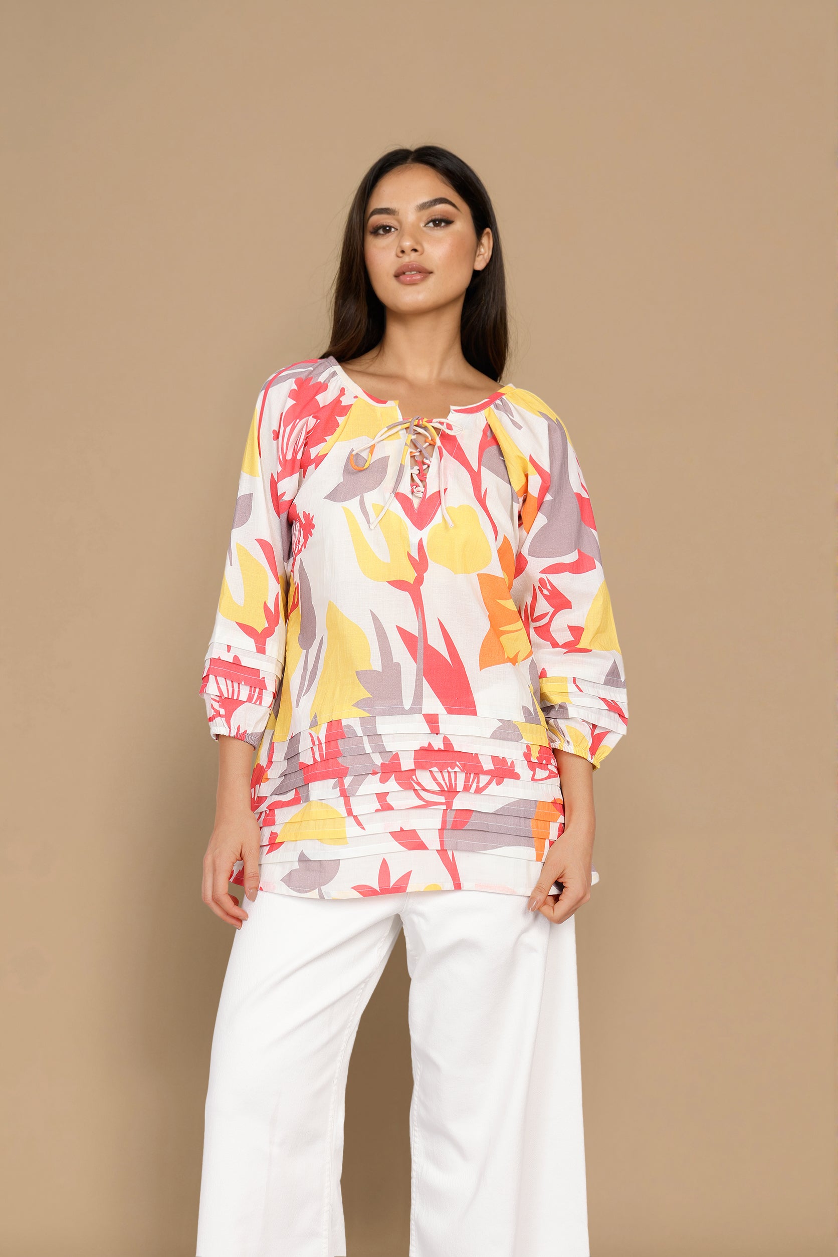 A woman modeling a three-quarter sleeve top with a colorful abstract floral print in yellow, red, and gray on a white base, paired with white wide-leg pants, set against a solid beige background.