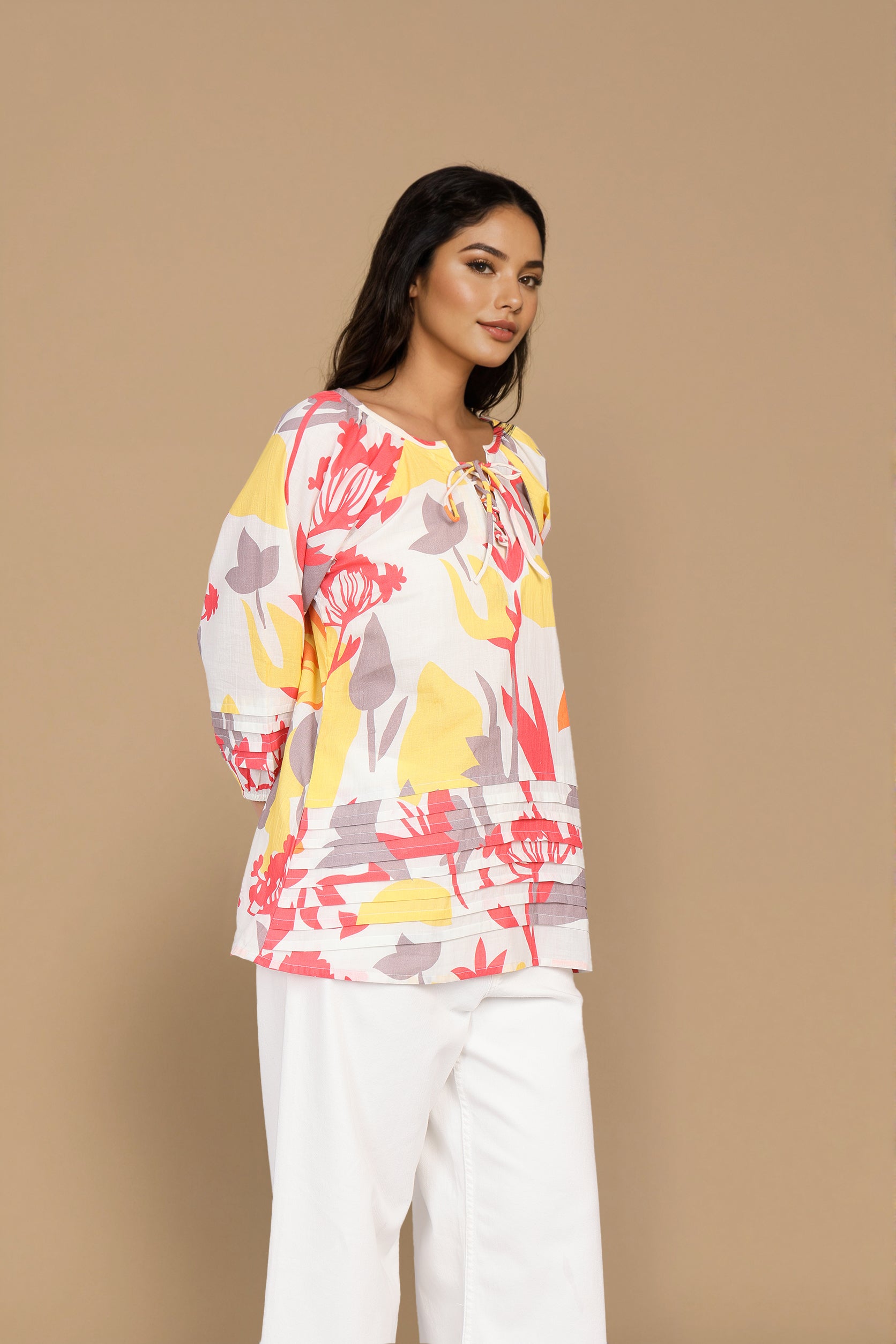 A woman modeling a long-sleeve top with a colorful floral pattern in pink, yellow, and gray on a white base, paired with white pants, against a solid beige studio background.