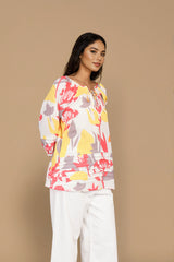 A woman modeling a long-sleeve top with a colorful floral pattern in pink, yellow, and gray on a white base, paired with white pants, against a solid beige studio background.