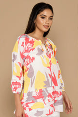 A woman modeling a white three-quarter sleeve tunic top with a vibrant tropical floral print in yellow, coral pink, and gray, set against a solid beige studio background.