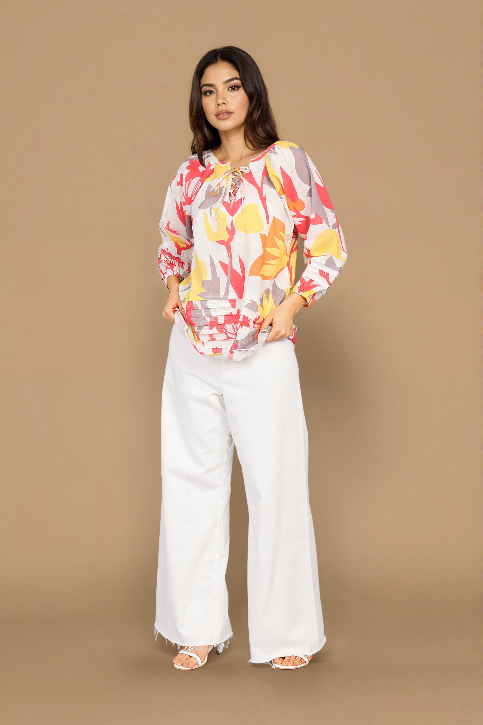 A woman modeling a long-sleeve blouse with a colorful floral and abstract print on a white base, paired with white wide-leg pants, against a solid tan studio background.