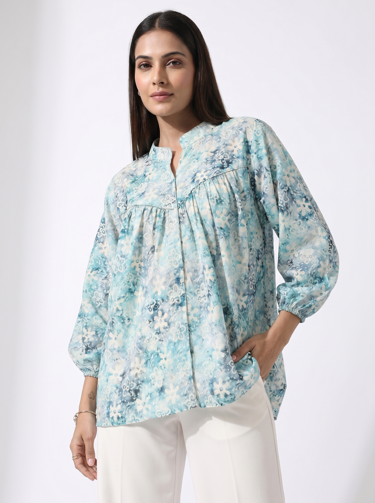 A woman modeling a light blue and white floral print blouse with a split neckline and gathered sleeves, paired with white pants, against a plain light gray studio background.