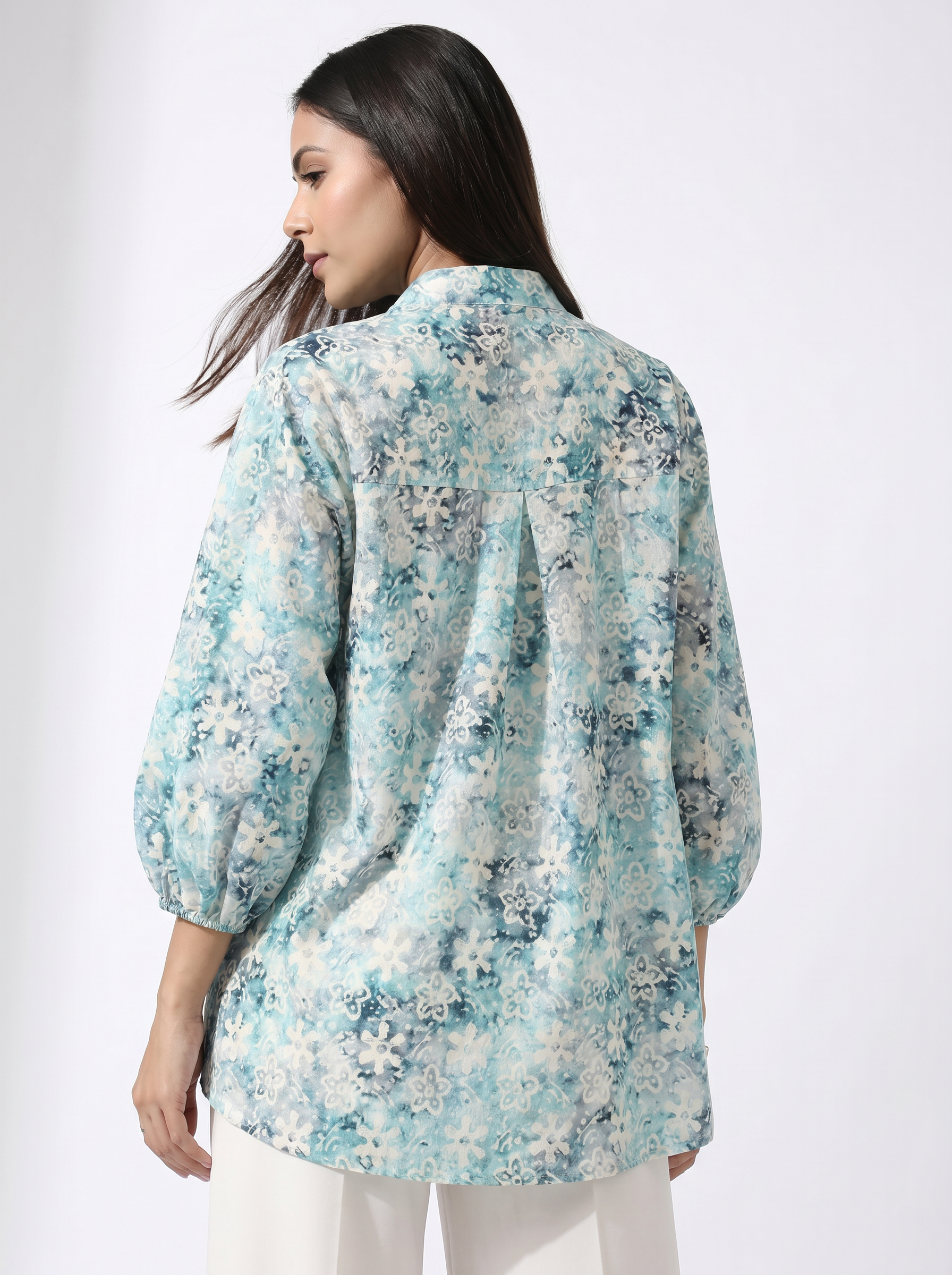 A woman shown from the back wearing a light blue and white floral print blouse with three-quarter sleeves, paired with white pants, against a plain white studio background.