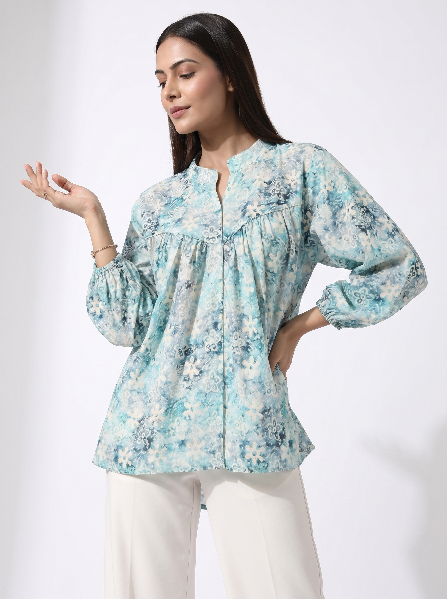A woman modeling a light blue and white floral print long-sleeve blouse with a split neckline, paired with white pants, against a plain white studio background.