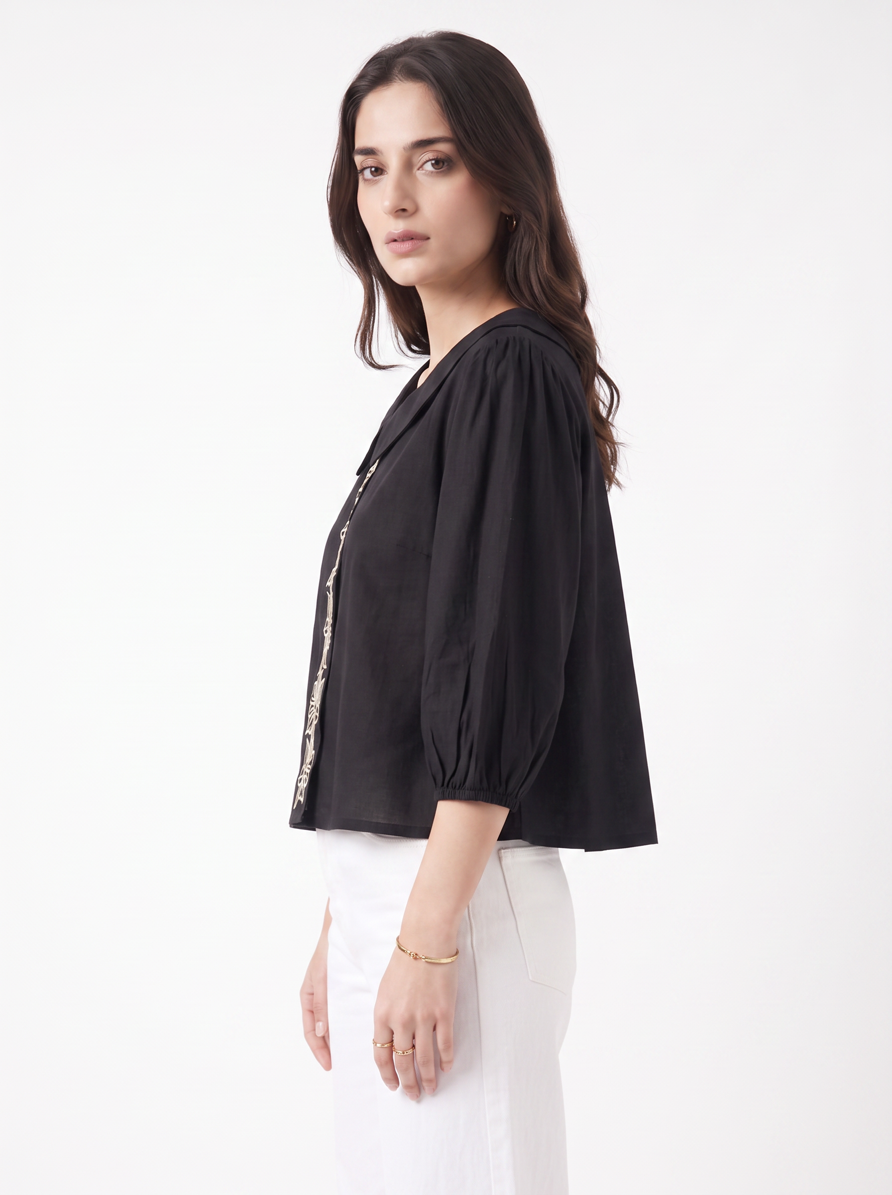 A woman wearing a black cropped puff-sleeve blouse with gold trim down the front, paired with white trousers, photographed against a plain white studio background.