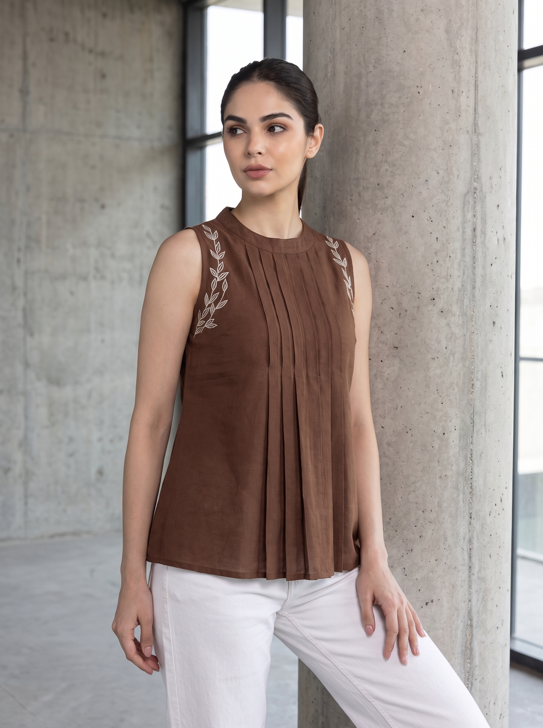 A woman wearing a sleeveless brown pleated top with embroidered details on the shoulders and white pants, posing in front of a concrete pillar.