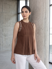 A woman wearing a sleeveless brown pleated top with embroidered details on the shoulders and white pants, posing in front of a concrete pillar.