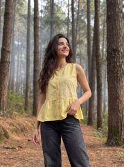 A woman wearing a sleeveless yellow top with white embroidery and dark gray jeans, standing on a forest path surrounded by tall trees.