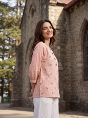 A woman wearing a light pink linen button-up top with small green floral embroidery and white pants, standing outdoors in front of a stone building.