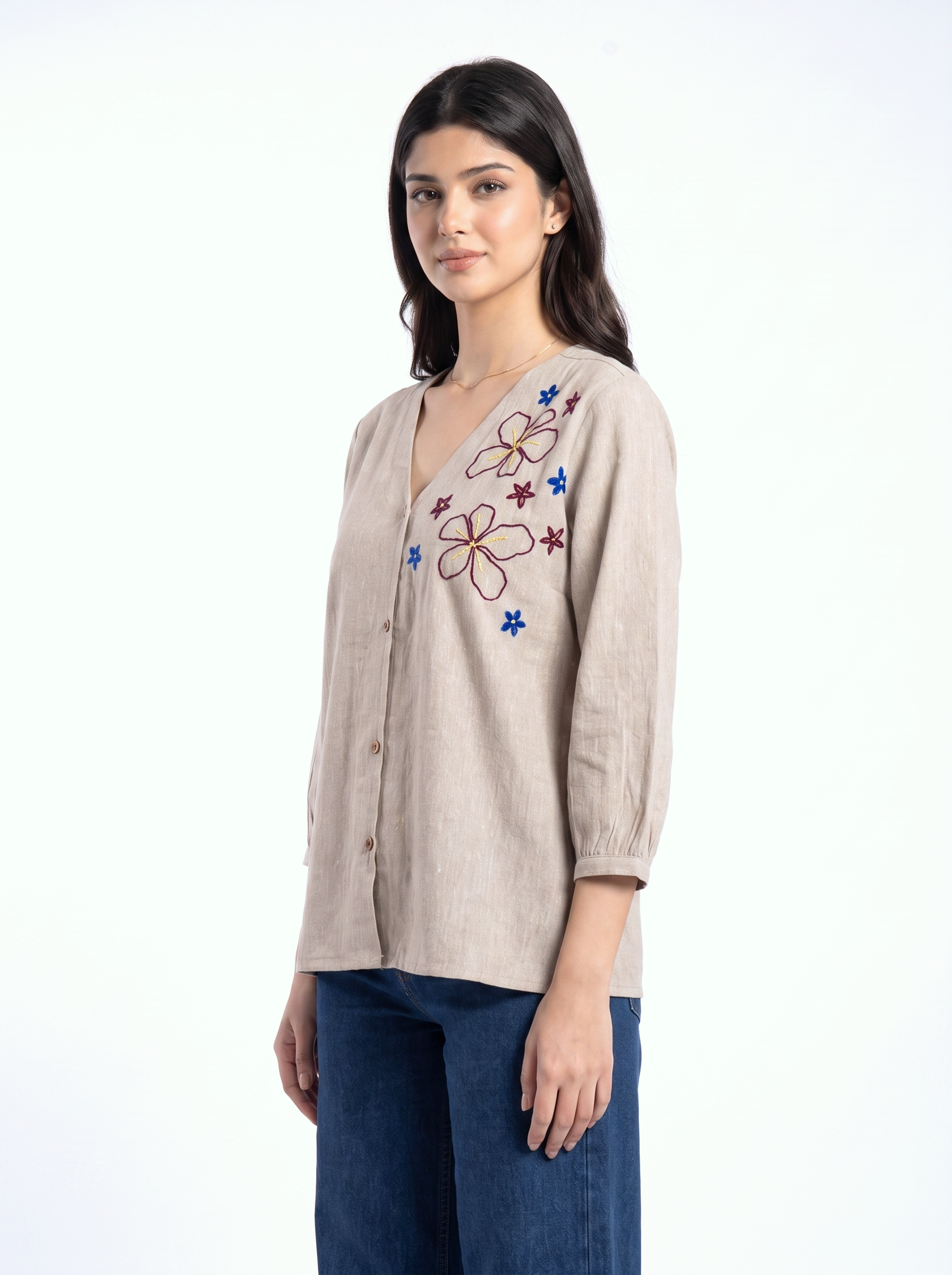 A woman modeling a beige linen button-up shirt with colorful floral embroidery on the chest, paired with dark blue jeans, against a plain white background.