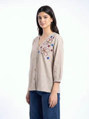 A woman modeling a beige linen button-up shirt with colorful floral embroidery on the chest, paired with dark blue jeans, against a plain white background.