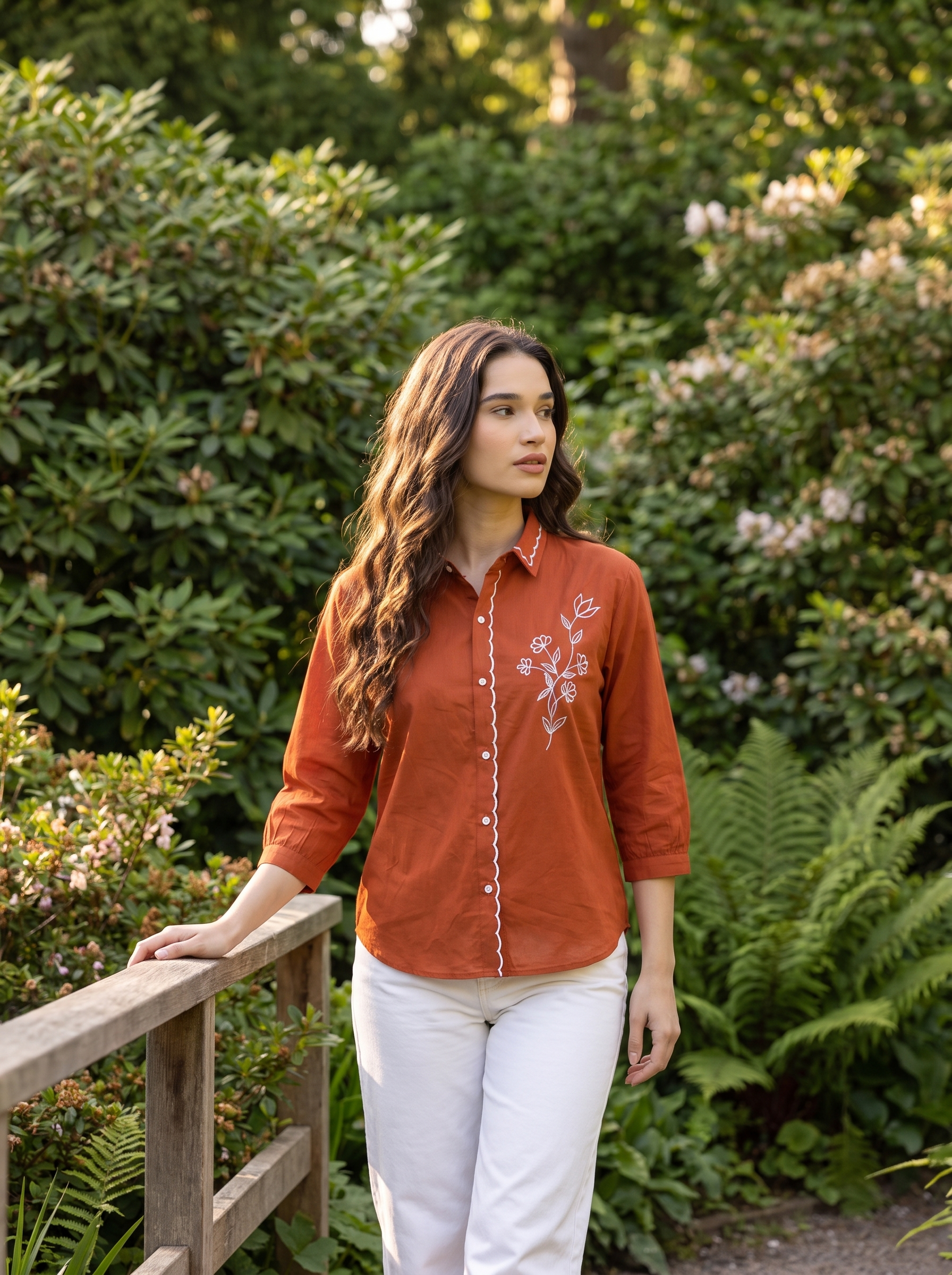 A woman wearing a rust-orange embroidered button-up shirt and white pants stands on a wooden bridge in a lush green garden.
