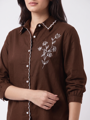 A close-up of a woman wearing a brown long-sleeve button-up shirt with white scalloped trim and white floral embroidery on the chest.