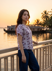 A woman stands by a railing at sunset beside a body of water, wearing a short-sleeve V-neck blouse with a purple and white floral pattern and dark blue jeans.